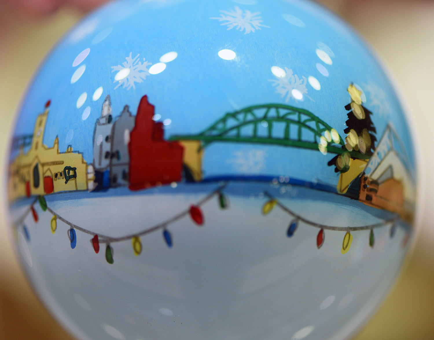 A close up photograph of a white and blue bauble with small painted Sunderland buildings and landmarks in a line across the middle with colourful string lights underneath and pale snowflakes above. From left to right: The Empire Theatre, Wearmouth Bridge, a large Christmas tree with a yellow star on top and the Stadium of Light.