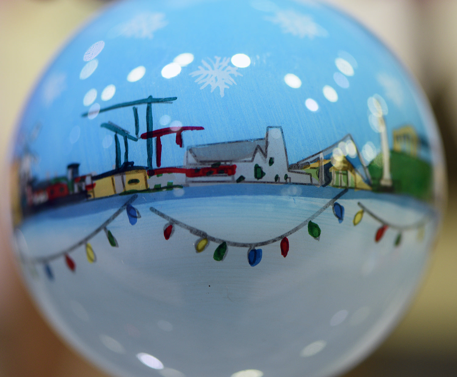 A close up photograph of a white and blue bauble with small painted Sunderland buildings and landmarks in a line across the middle with colourful string lights underneath and pale snowflakes above.