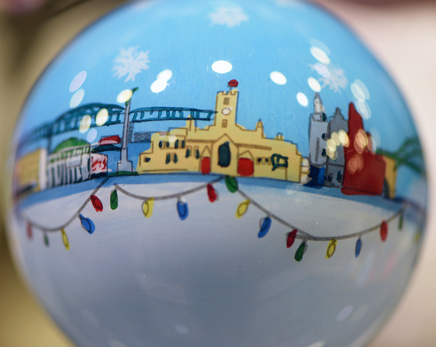 A close up photograph of a white and blue bauble with small painted Sunderland buildings and landmarks in a line across the middle with colourful string lights underneath and pale snowflakes above. From left to right: Sunderland Museum and Winter Gardens, Mowbray Park, Alexandra Bridge, a castle, The Empire Theatre and Wearmouth Bridge.