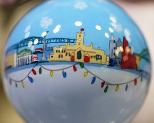 A close up photograph of a white and blue bauble with small painted Sunderland buildings and landmarks in a line across the middle with colourful string lights underneath and pale snowflakes above. From left to right: Sunderland Museum and Winter Gardens, Mowbray Park, Alexandra Bridge, a castle, The Empire Theatre and Wearmouth Bridge.