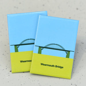 Two flat, rectangle magnets sat one on top of the other - they feature a graphic illustration of an arched bridge in shades of green against a blue background. Below the bridge is a section of solid light green, inside this section, dark green bold text reads "Wearmouth Bridge"
