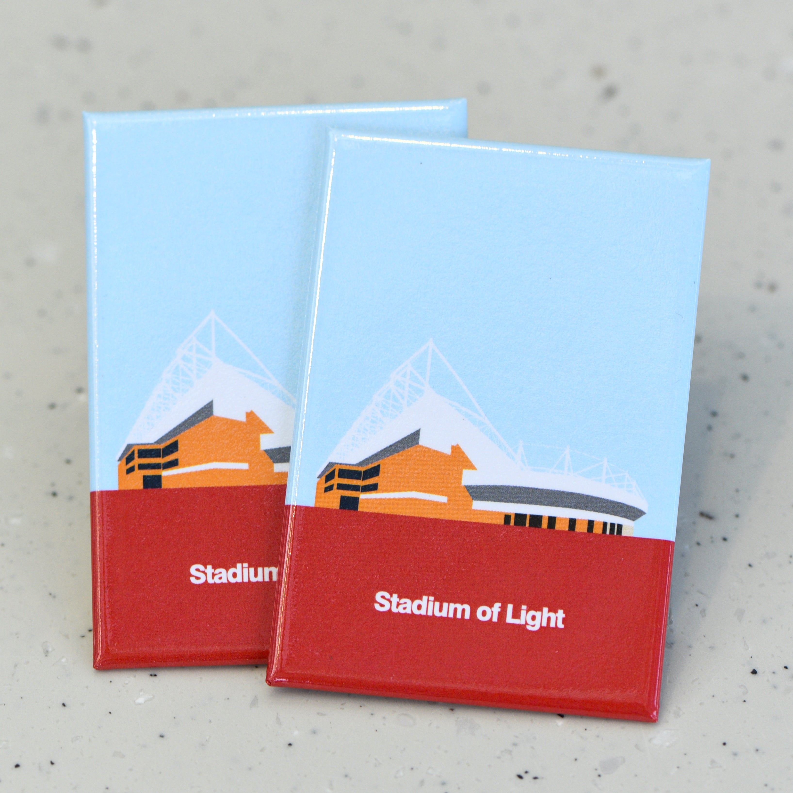 Two flat rectangle magnets featuring a colourful graphic illustration of a football stadium in orange, grey and white against a pale blue sky. A large section of solid red at the bottom of the magnet contains white bold text that reads "Stadium of Light".