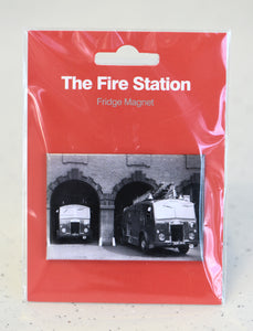 A flat, landscape, rectangle magnet featuring a black and white photograph of two fire engines emerging from decorated archways side by side. The fire engine on the left is only partially visible whereas the one on the right is almost completely out of the archway. The magnet sits on red card wrapped in plastic, at the top of the card, bold white text reads "The Fire Station"