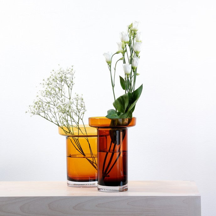 Two glass vases with a cylinder shape that widens into a wide flat cylinder at the top sit side by side - the vase to the left is amber and sits shorter and wider, the vase to the left is taller and narrower and brown. Both hold green and white foliage.