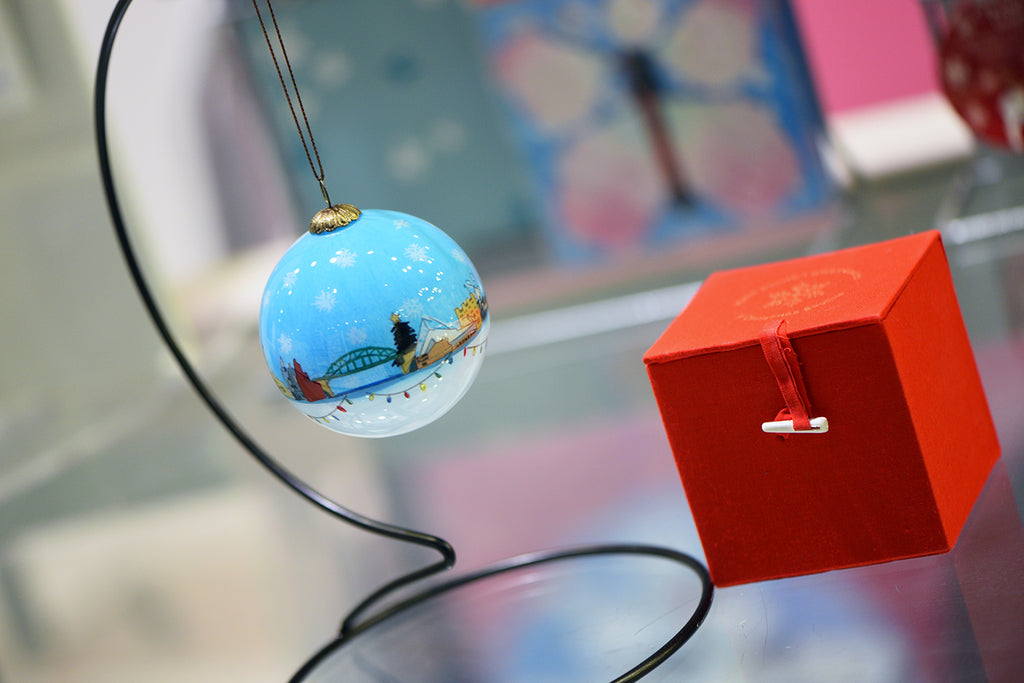 A glass bauble painted white and blue featuring a line of hand painted Sunderland landmarks and buildings around the middle and colourful string lights underneath. The bauble hangs on a black hanger next to a red fabric gift box.